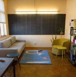 photo of the office - windows on the left, blackboard in the back, warm wooden floors with a blue rug featuring an abstract pattern in orange, light pink, and black. there is a grey rectangular couch on the left and a small, curved green armchair on the right.